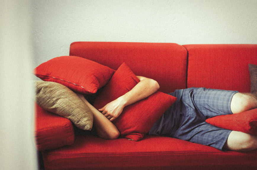 A man lies on a couch hiding his head with cushions as he is experiencing severe untreated food intolerance