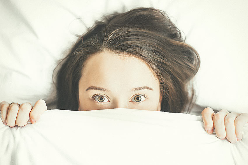 A young woman peeks out from her bed sheets for a blog-post on sleep