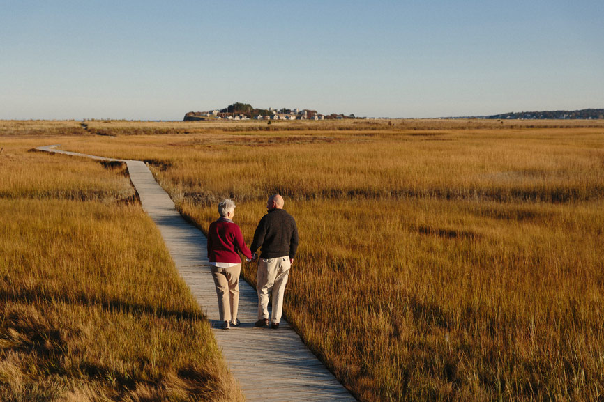 An elderly couple hold hands as they walk up a path amongst dry grass